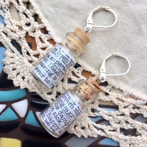 May include: A pair of silver earrings with small glass bottles attached. The bottles have cork stoppers and contain handwritten notes. The notes in the bottles read "I've been trying to drown my daughter in the damn bathtub. Not sick, I am happy about it. De..." and "I have been trying to drown my daughter in the damn bathtub. Not sick, I am happy about it. De..."
