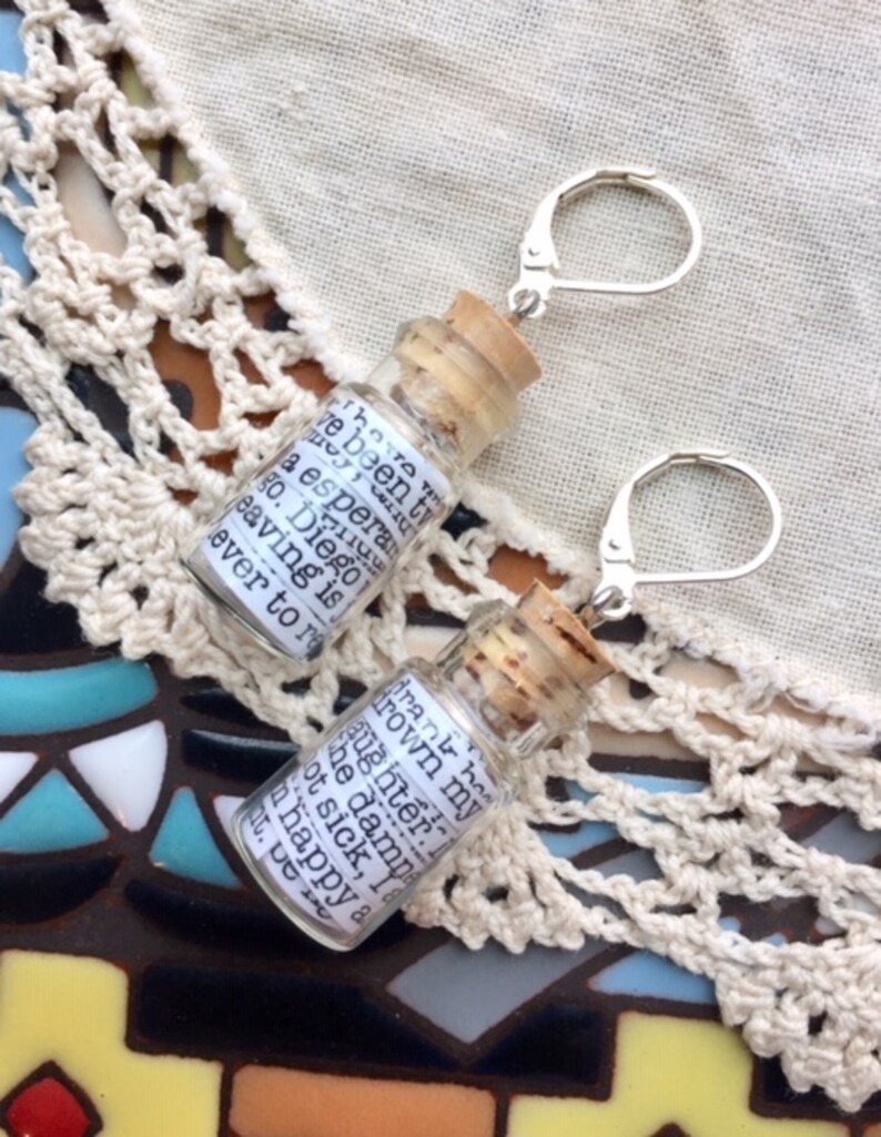 May include: A pair of silver earrings with small glass bottles attached. The bottles have cork stoppers and contain handwritten notes. The notes in the bottles read "I've been trying to drown my daughter in the damn bathtub. Not sick, I am happy about it. De..." and "I have been trying to drown my daughter in the damn bathtub. Not sick, I am happy about it. De..."