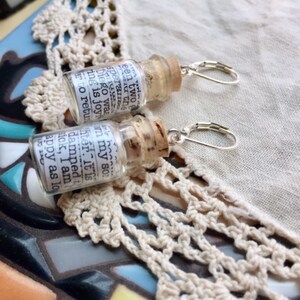 May include: A pair of silver earring hooks with small glass bottles attached. The bottles are filled with a white paper with text that reads: "been two speraira Diego was ing is joy in my sor ter. It is damned t sick, I am appy as lo 0120"