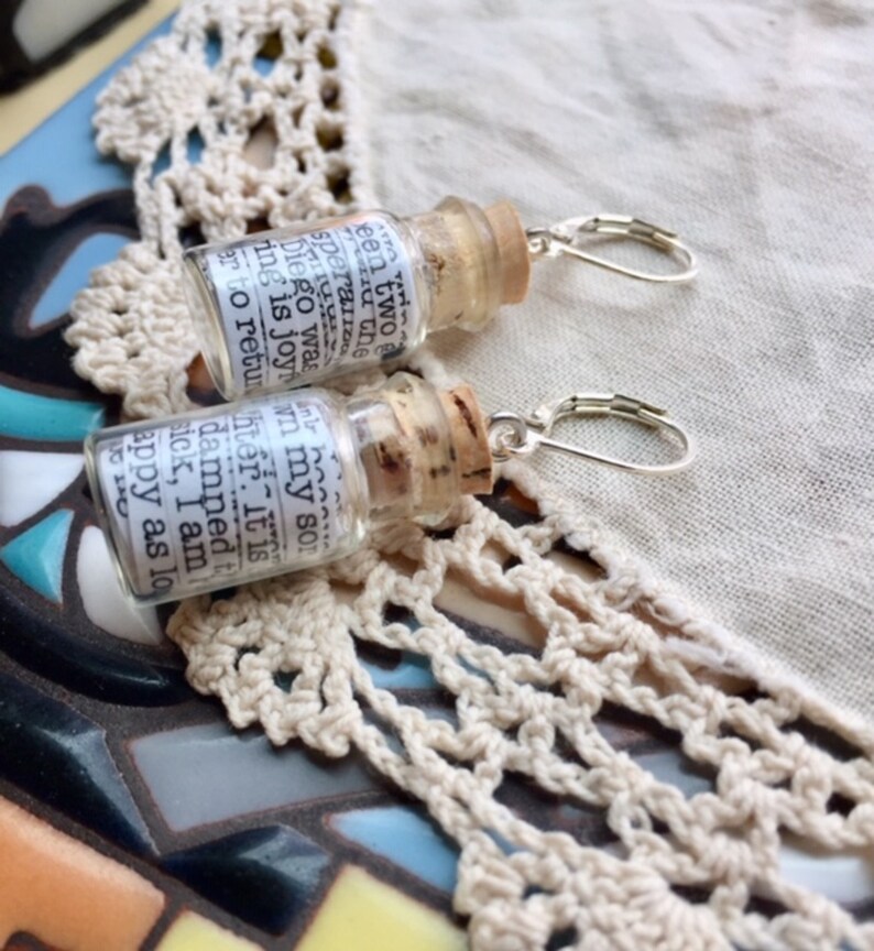 May include: A pair of silver earring hooks with small glass bottles attached. The bottles are filled with a white paper with text that reads: "been two speraira Diego was ing is joy in my sor ter. It is damned t sick, I am appy as lo 0120"