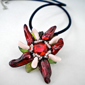 Puede incluir: Un collar con un colgante de flor roja y blanca con una gran piedra preciosa roja en el centro. El colgante está hecho de cuentas y tiene un cordón negro.