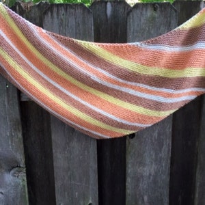 May include: A striped knitted scarf in shades of brown, orange, yellow, and white. The scarf is draped over a wooden fence.