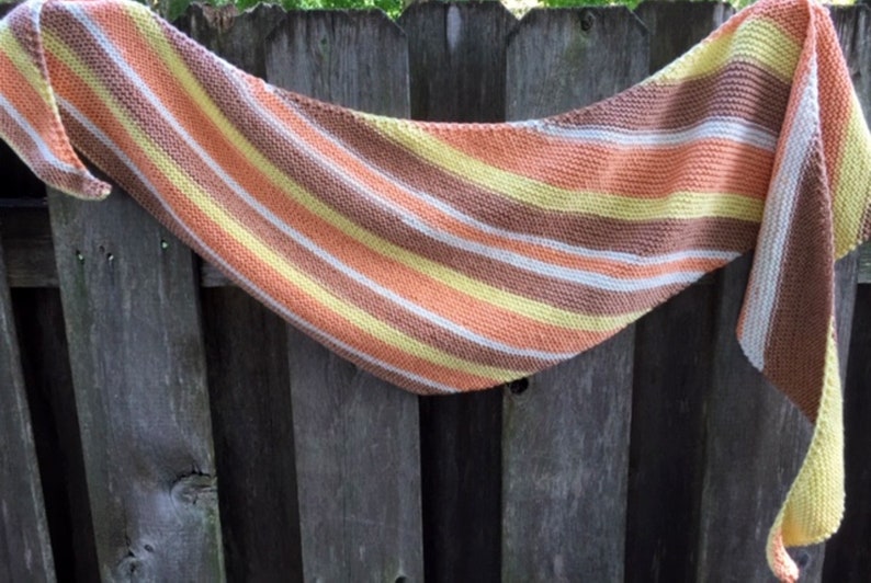 May include: A striped knitted scarf in shades of brown, orange, yellow, and white. The scarf is draped over a wooden fence.