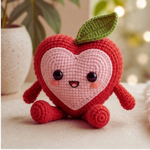 Può includere: Un giocattolo di peluche a forma di cuore fatto a mano all'uncinetto. Il giocattolo è principalmente rosso con un cuore rosa al centro. Ha occhi neri, una bocca sorridente e una foglia verde. Il giocattolo ha braccia e gambe piccole.