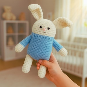 May include: A handmade crochet bunny doll with a white head and limbs, and a light blue body. The bunny has black eyes and a pink nose. The doll is being held in a person's hand. The background is blurred, showing a crib and other objects.