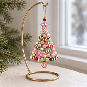 Könnte beinhalten: Ein kegelförmiger Weihnachtsbaumschmuck aus verschiedenen Perlen in Rosa, Grün, Weiß und Rot. Er ist mit Miniatur-Zuckerstangen und Lutscher-Akzenten verziert, wird von einem rosa Stern gekrönt und hängt an einem goldfarbenen Metallständer.