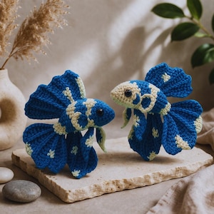 Op de afbeelding: Twee handgemaakte gehaakte betta-vissen in blauw en crème. De vissen hebben gedetailleerde vinnen en staarten, met crèmekleurige accenten en donkere ogen. Ze worden gepresenteerd op een stenen oppervlak, wat duidt op een decoratief of artistiek thema.