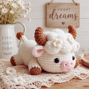 Peut inclure: Une peluche au crochet en forme de vache avec un corps blanc, des cornes brunes, des sabots bruns et un museau rose. La vache présente une tête blanche duveteuse avec de petites fleurs blanches, des yeux noirs et des oreilles roses, sur un napperon.