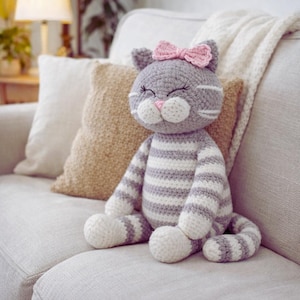 Könnte beinhalten: Gehäkelte Katzendoll mit grau-weißen Streifen, einer rosa Schleife und geschlossenen Augen. Die Katze hat weiße Pfoten und eine rosa Nase. Sie sitzt auf einem hellen Sofa mit Kissen im Hintergrund.