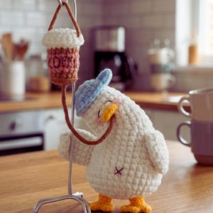Puede incluir: Una caprichosa figura de pato de ganchillo hecha a mano con gorra azul y pies amarillos, sosteniendo una taza de café en miniatura. La taza de café es marrón y blanca, con la palabra "COFFEE" visible. El pato es blanco con una pequeña cruz negra en la parte trasera.