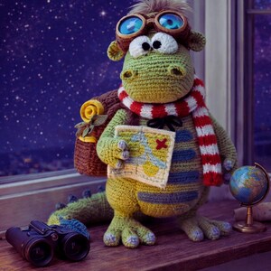 Peut inclure: Une figurine de dragon au crochet, portant des lunettes, une &eacute;charpe et un sac &agrave; dos, tenant une carte. Un petit globe et des jumelles sont &agrave; proximit&eacute;. Le dragon est vert, jaune et bleu, avec une &eacute;charpe ray&eacute;e rouge et blanche. Le fond est un ciel &eacute;toil&eacute;.