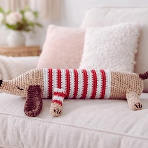 Peut inclure: Un coussin en forme de chien teckel crocheté, de couleur beige, avec un pull rayé rouge et blanc. Le chien a un nez noir, des oreilles marron et une queue marron. Il est sur un canapé blanc.