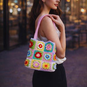 Könnte beinhalten: Eine farbenfrohe Häkeltasche mit rosa Trägern. Die Tasche hat ein Patchwork-Design aus quadratischen Blumenmotiven in verschiedenen Farben, darunter Rot, Gelb, Blau und Grün. Die Tasche wird von einer Person gehalten.