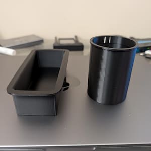 May include: Two black 3D-printed plastic containers. One is rectangular with a lip, the other is cylindrical with vertical slits near the top. Both are on a gray surface.