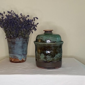 May include: A cylindrical ceramic vase filled with dried purple lavender stems next to a lidded ceramic jar. The vase features a blue and brown glaze, while the jar has a dark brown base transitioning into a mottled green and black glazed lid.