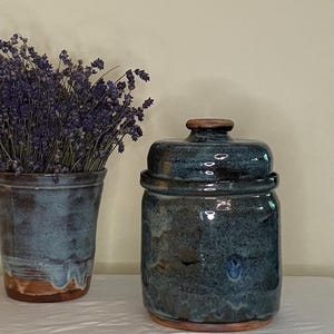 May include: A cylindrical ceramic canister with a lid, featuring a mottled blue and brown glaze. Beside it stands a matching ceramic vessel filled with dried purple lavender sprigs. Both items exhibit a glossy finish and exposed clay base.