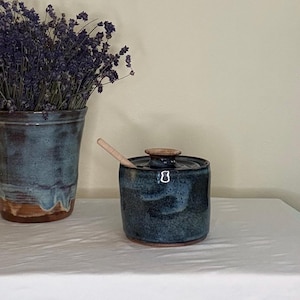 May include: A cylindrical ceramic honey pot with a dark blue and black speckled glaze, featuring a matching lid and a light-colored wooden honey dipper resting inside. Beside it stands a taller, matching ceramic vase filled with dried lavender.