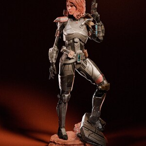 May include: A detailed figurine of a woman in futuristic armor, holding a gun. The figure is predominantly silver and black, with red accents on the shoulders and knee. The woman has red hair and is posed on a reddish-brown rock.