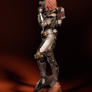 May include: A detailed figurine of a female character in futuristic armor, holding a gun. The figure has red hair and is posed on a rocky base. The armor is primarily silver and black with gold and pink accents. A backpack is visible on the back.