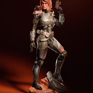 May include: A detailed figurine of a female character in futuristic armor, holding a gun. The figure is posed on a rocky base, with a dark red background. The armor is primarily gray with red and blue accents. The character has short, reddish-brown hair.