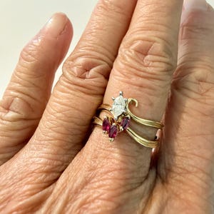 May include: A gold ring set featuring a marquise-cut diamond and three ruby accents. The ring has a unique design with a curved band and is set on a finger. The ring is a piece of jewelry.