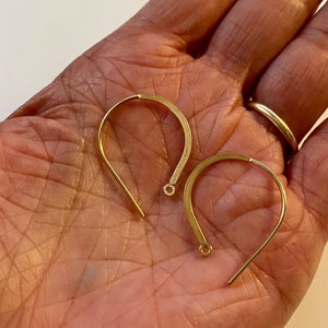 May include: Two gold-toned earring hooks, each with a small loop at the end. The hooks are lying on a person's hand.