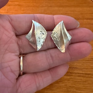 May include: A pair of silver-toned earrings. Each earring has a geometric, diamond-like shape with a floral design etched into the surface. The earrings are held in a hand, showcasing their size and design.