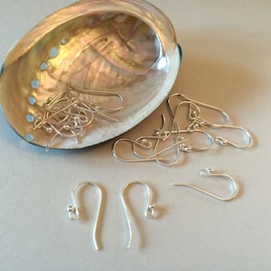 May include: Silver-colored earring hooks and components are scattered on a light gray surface, with some pieces inside a pearlescent seashell. The hooks are curved with a small ball at the end, ideal for jewelry making.