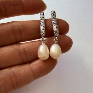May include: A pair of drop earrings featuring elongated silver posts with a textured, repeating oval pattern. Each post connects to a smooth, cream-colored teardrop-shaped pearl that hangs vertically from a small silver jump ring at the base.