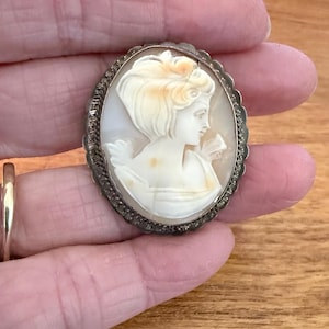 May include: Oval cameo brooch featuring a profile of a woman with flowing hair and a delicate floral detail. The cameo is cream-colored with light brown accents, set in a silver-toned, ornate frame. The brooch is held in a hand.