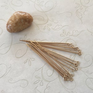 May include: A group of 15 gold head pins with loops. The head pins are arranged in a fan shape on a white surface with a floral pattern.