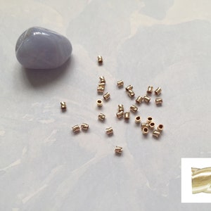 May include: A scattering of gold-colored metal crimp beads on a light gray surface. A single, light blue, smooth, oval-shaped stone is in the upper left corner of the image. A close-up of a single crimp bead is in the lower right corner of the image.