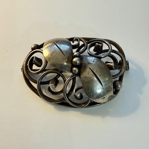 May include: An antique silver-toned brooch with an oval shape. The brooch features two stylized leaf shapes and a decorative openwork design with swirling lines and small spheres. The brooch has a pin clasp on the back.