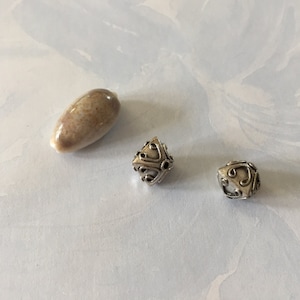 May include: An assortment of beads and a polished stone. The stone is oval-shaped with a speckled tan and brown pattern. Two silver-colored beads with intricate designs are also present, one with a geometric pattern and the other with a swirling design.
