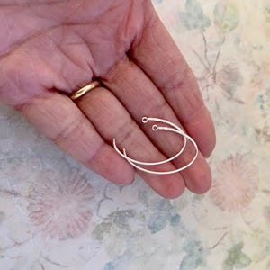 May include: A pair of silver hoop earrings with a delicate, curved design. The earrings are held in a hand, with a gold ring visible on a finger. The background features a floral pattern.