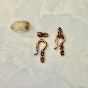 May include: A collection of copper-colored jewelry clasps and a small, oval-shaped, beige stone. The clasps include a hook clasp, a small connector, and a decorative clasp with leaf-like details. The items are arranged on a light blue and white surface.
