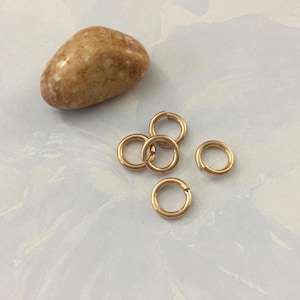 May include: Five small, circular, gold-toned metal jump rings with a visible split in each ring, arranged in a cluster next to a single, smooth, tan and white speckled river stone with a polished, rounded surface texture.