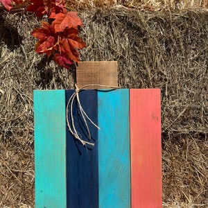 May include: A wooden pumpkin decoration painted in teal, navy blue and coral. The pumpkin is made from four individual planks of wood and is sitting on a bale of hay.
