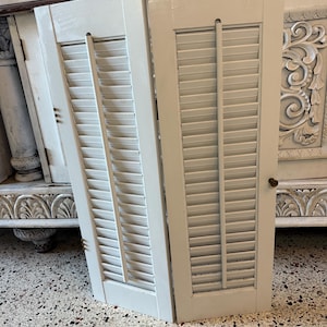 May include: Two off-white wooden shutters with horizontal slats and a vertical support. The shutters are leaning against a carved wooden cabinet. The shutters have a simple, classic design and are painted in a neutral color.