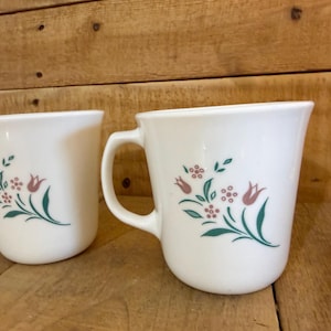 May include: Two white milk glass mugs featuring a floral pattern of pink tulips and small pink blossoms with green leaves. Each mug has a curved handle and a smooth, rounded body, showcasing a consistent botanical design on the front surface.