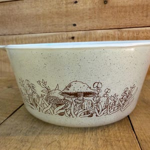 May include: A cream-colored mixing bowl with a speckled finish and a brown line art illustration of mushrooms and foliage wrapping around the exterior. The bowl features a wide rim and a small handle tab on the right side for easy gripping.