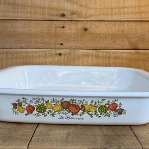 May include: A rectangular white enamel baking dish featuring a colorful botanical illustration of various fruits and vegetables along the front side, with the cursive text Le Romarin printed in black ink below the produce design.