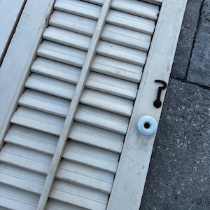 May include: White wooden shutters with a rusty metal hook and a white plastic knob. The shutters are closed and have a weathered look.