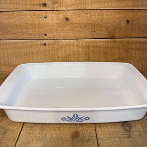 May include: A rectangular white ceramic baking dish with rounded corners and a smooth, glossy finish. The exterior front side features a small, centered blue floral pattern consisting of stylized flowers and leaves arranged in a symmetrical design.