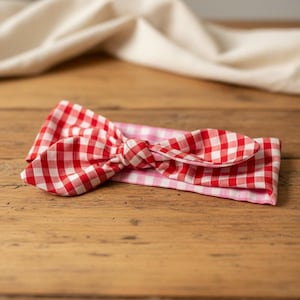 Puede incluir: Una diadema con estampado de cuadros rojos y blancos con un detalle de lazo anudado. La diadema es de tela y est&aacute; sobre una superficie de madera. Un pa&ntilde;o blanco en el fondo.