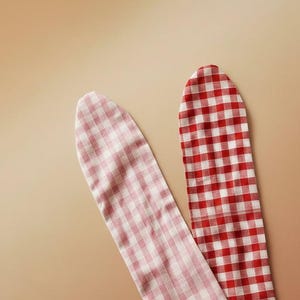Puede incluir: Dos tiras de tela con estampado de cuadros vichy. Una tira es roja y blanca, la otra rosa y blanca. Las tiras est&aacute;n colocadas en diagonal sobre un fondo beige. La tela parece ser de algod&oacute;n o un material similar.