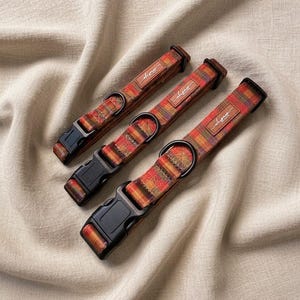 May include: Three pet collars featuring a multicoloured plaid pattern in shades of orange, red, yellow, and purple. Each collar includes a black plastic side-release buckle, a black metal D-ring, and a small brown rectangular leather-like brand tag.