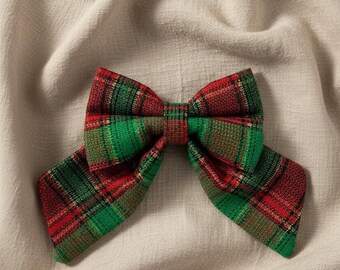 May include: A fabric hair bow featuring a classic plaid pattern in vibrant shades of red, forest green, and black. The bow is constructed with a central knot and two wide, angled tails extending outward, showcasing a textured, woven material finish.
