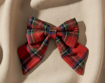 May include: A hair bow featuring a classic red, green, and blue plaid tartan pattern. The bow is constructed with two main loops and two pointed tails extending downward, showcasing a structured fabric design with a centered knot detail.