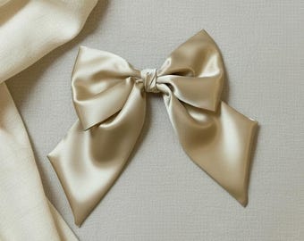 May include: A champagne-colored satin hair bow featuring a smooth, lustrous finish with two wide, pointed tails extending downwards. The bow is tied in the center with a small knot, creating a structured shape with soft folds and a reflective surface.